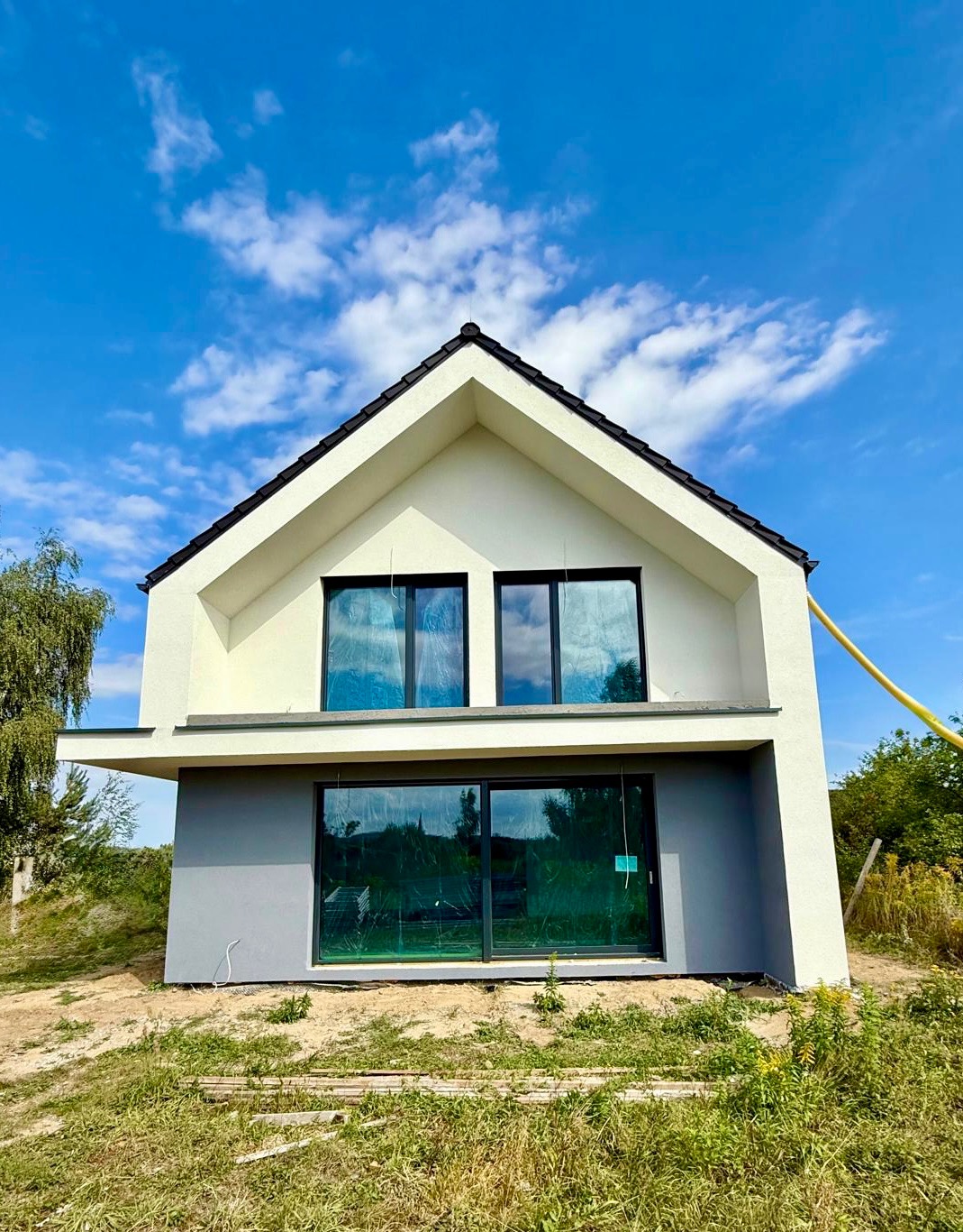 Nowoczesny dom jednorodzinny z dwukolorową elewacją (biały tynk i szary dół), duże okna zabezpieczone folią, błękitne niebo w tle. Widok z dołu.