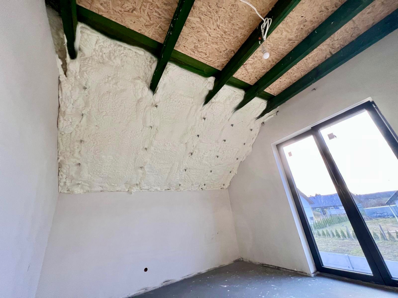 Poddasze z widoczną izolacją pianką PUR między krokwiami, surowy stan ścian, okno dachowe z widokiem na zewnątrz. Detal: widoczne mocowania pianki.