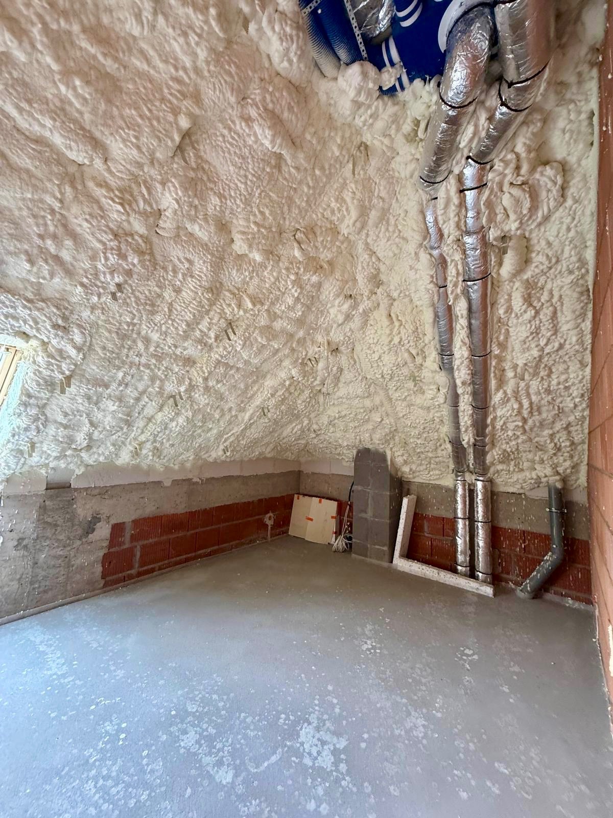 Poddasze po ociepleniu pianką PUR, widoczne rury wentylacyjne w srebrnej izolacji. Ściany częściowo wykończone cegłą, surowy beton na podłodze.