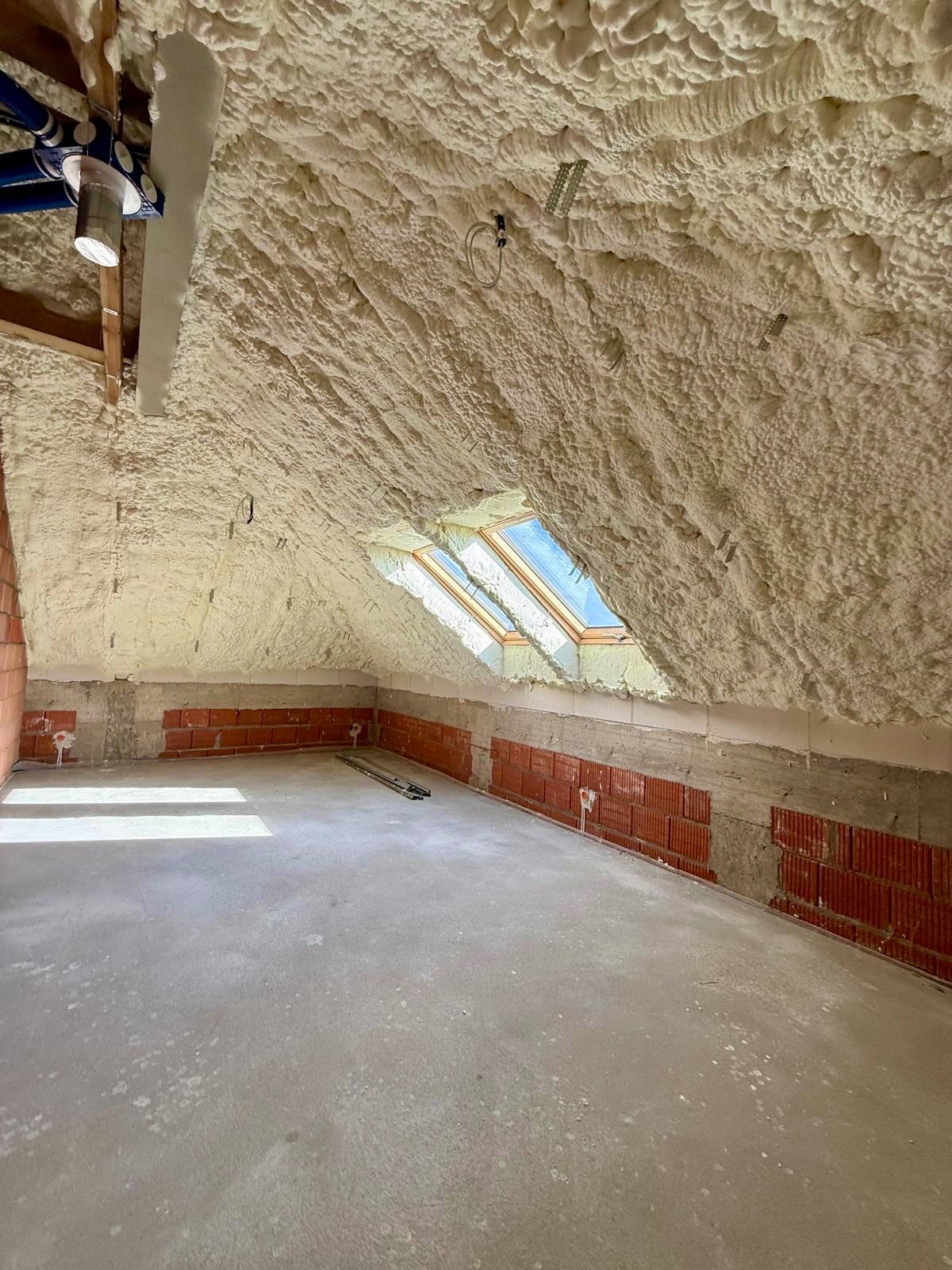 Poddasze po ociepleniu pianką PUR, widoczne okna dachowe i instalacje. Ściany z cegły częściowo ocieplone. Szara, betonowa podłoga. Oświetlenie naturalne.