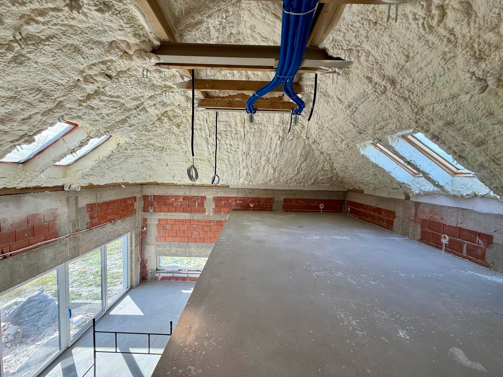 Poddasze po ociepleniu pianką PUR, widoczne okna dachowe i niebieskie rury wentylacyjne. Ściany z czerwonej cegły częściowo otynkowane. Surowe wykończenie.