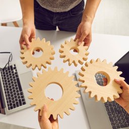 Agencja Interaktywna Sucharscy Karol Sucharski - Cztery drewniane koła zębate trzymane nad biurkiem z dwoma laptopami i okularami, symbolizujące współpracę i procesy.