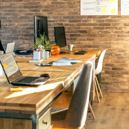 Agencja Interaktywna Sucharscy Karol Sucharski - Biuro z długim, drewnianym stołem, na którym znajdują się laptopy, monitory, notatki, artykuły piśmiennicze, rośliny doniczkowe i piłka do koszykówki; na ścianie z cegły wisi tablica z harmonogramem.