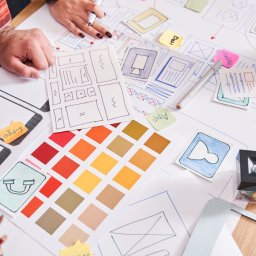 Agencja Interaktywna Sucharscy Karol Sucharski - Ręcznie rysowane szkice interfejsu aplikacji mobilnej, paleta kolorów i notatki na biurku podczas procesu projektowania.
