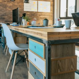 Agencja Interaktywna Sucharscy Karol Sucharski - Biuro w stylu loft: drewniany stół z szufladami o geometrycznych wzorach, białe krzesło, ceglana ściana, monitory i notatki na tablicy w tle.