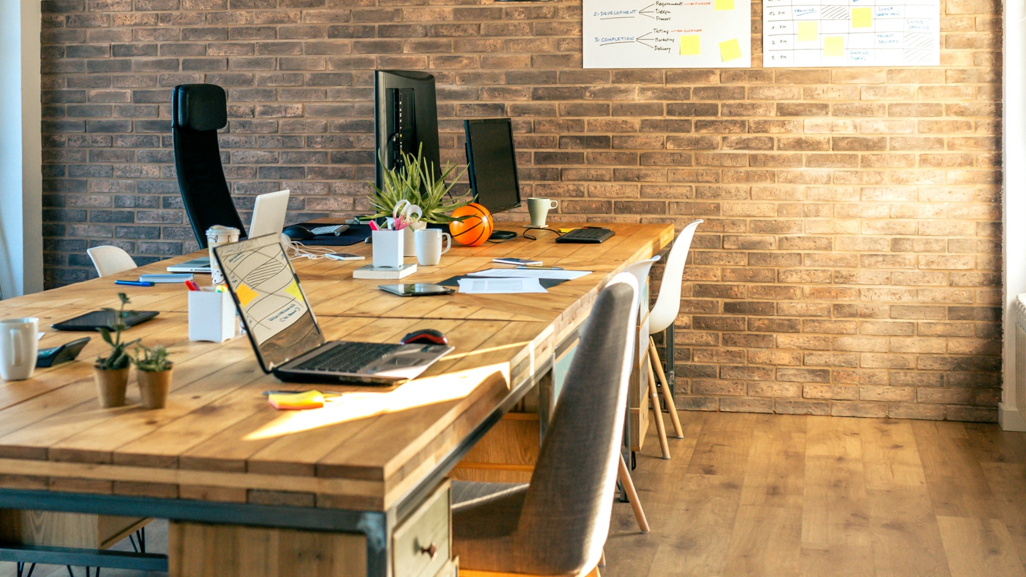 Biuro z długim, drewnianym stołem, na którym znajdują się laptopy, monitory, notatki, artykuły piśmiennicze, rośliny doniczkowe i piłka do koszykówki; na ścianie z cegły wisi tablica z harmonogramem.