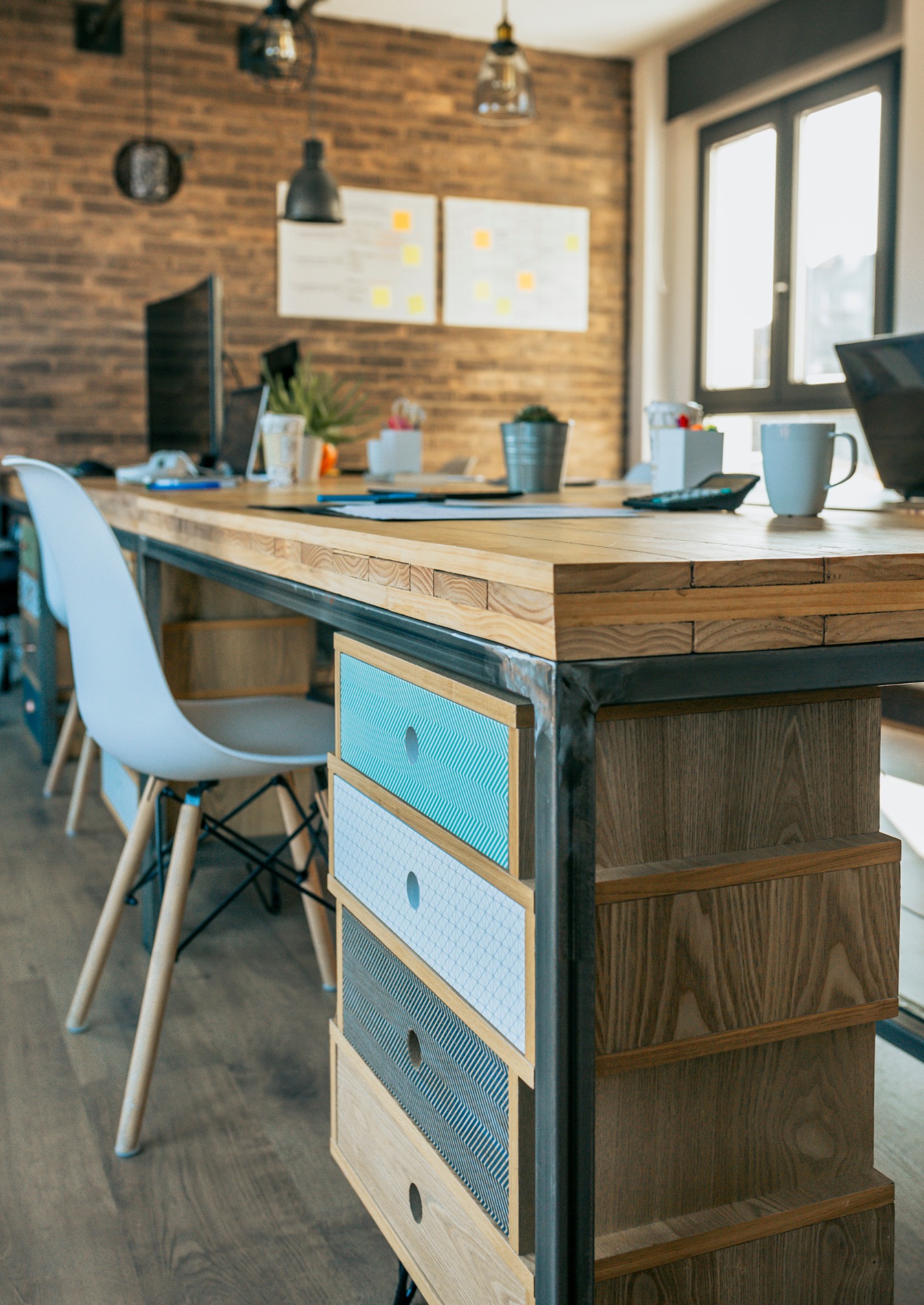 Biuro w stylu loft: drewniany stół z szufladami o geometrycznych wzorach, białe krzesło, ceglana ściana, monitory i notatki na tablicy w tle.