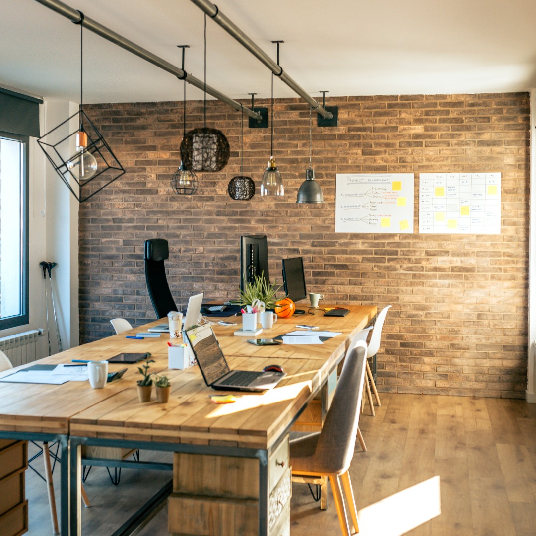 Przestrzeń biurowa z długim, drewnianym stołem, laptopami, monitorami, tablicami suchościeralnymi z notatkami i kalendarzem na ceglanej ścianie, oświetlona wiszącymi lampami.