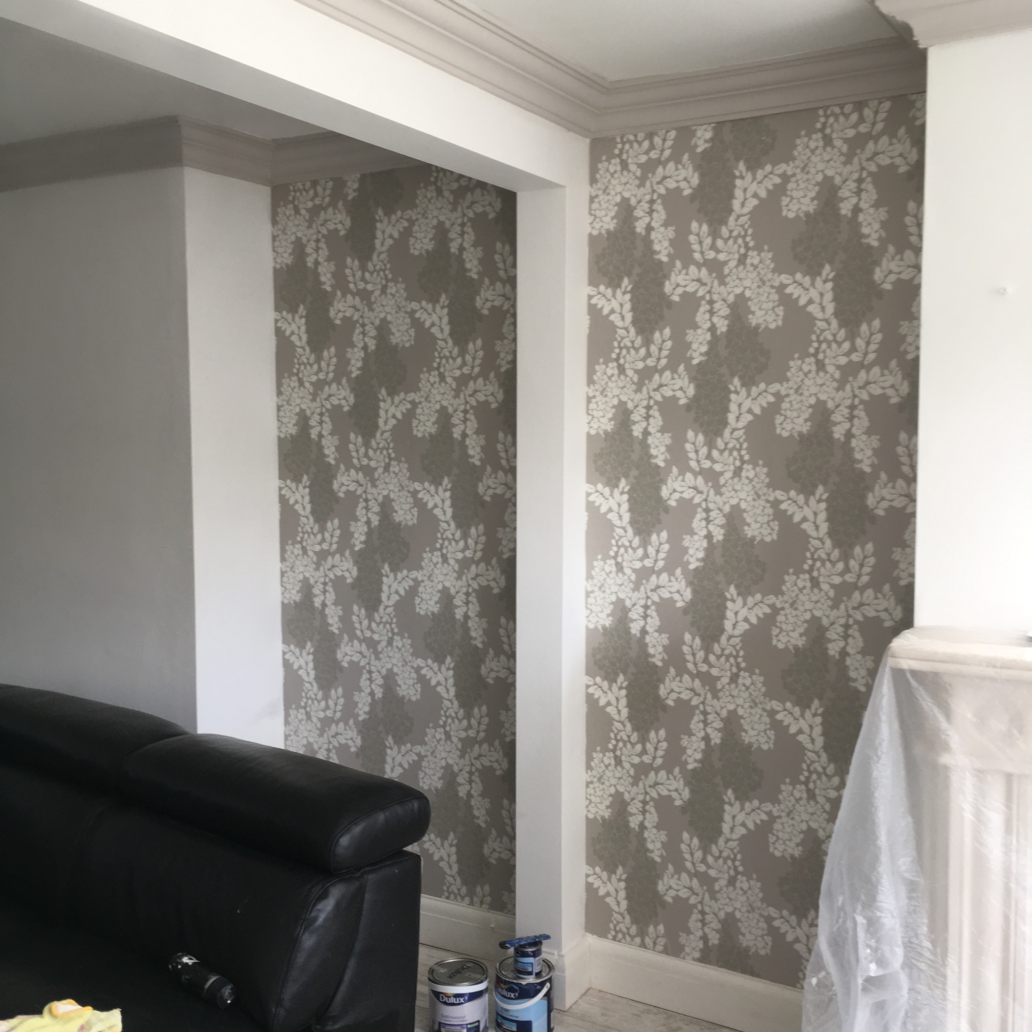 Pokój po tapetowaniu z wzorem liści, widoczny fragment czarnej sofy i puszki farby Dulux.