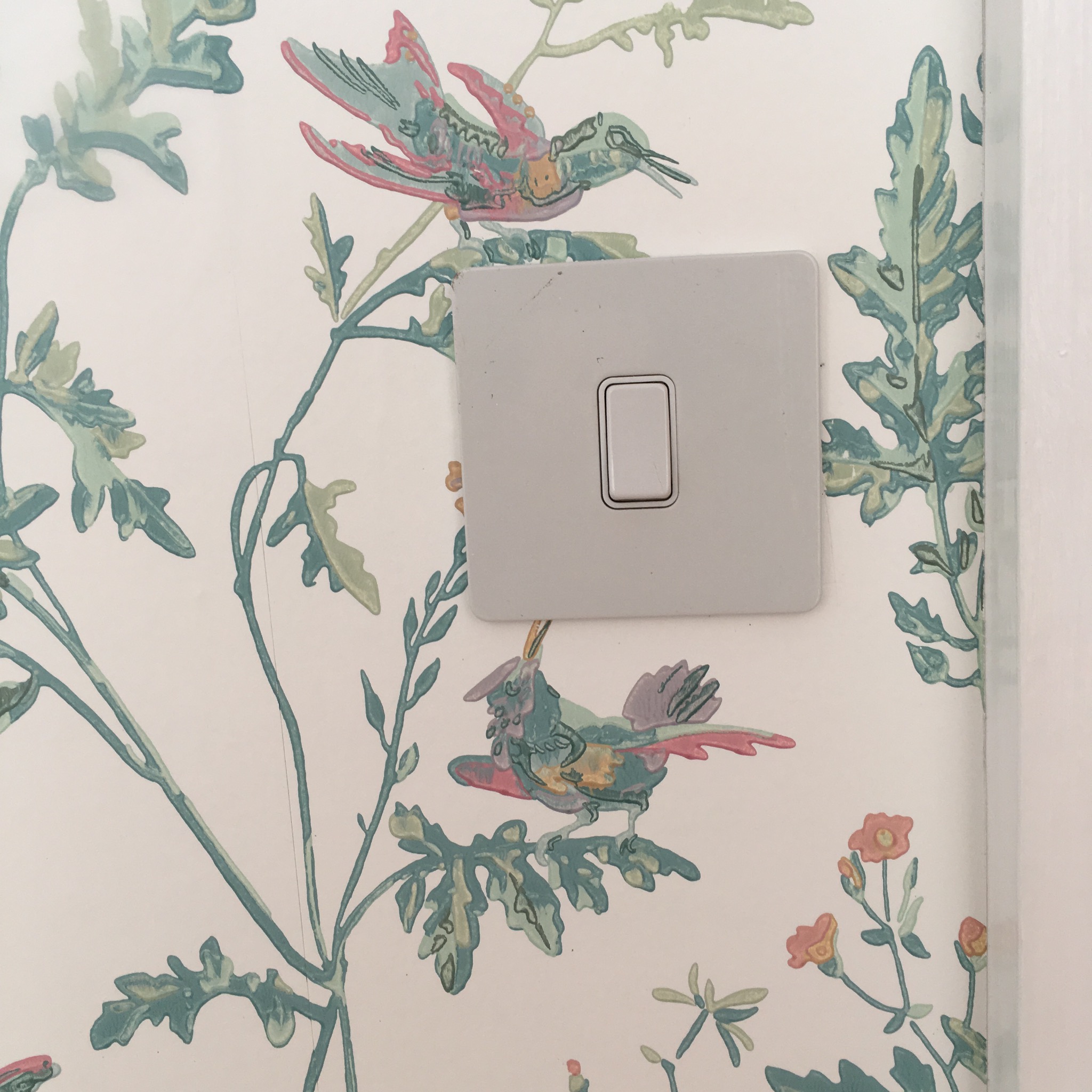 Fragment ściany z tapetą w delikatny wzór ptaków i roślin w pastelowych kolorach, obok włącznik światła z drobnymi zabrudzeniami.