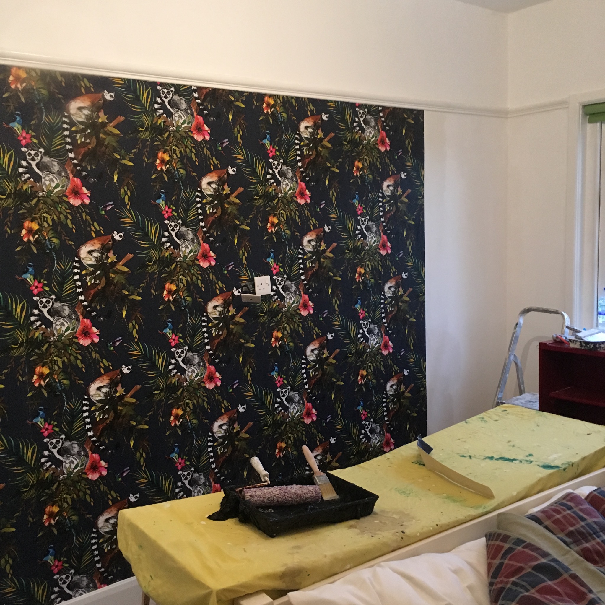 Ściana po nałożeniu tapety z motywem lemurów i tropikalnych kwiatów; widoczna rolka i pędzel malarski w kuwecie na zabezpieczonym podłożu.