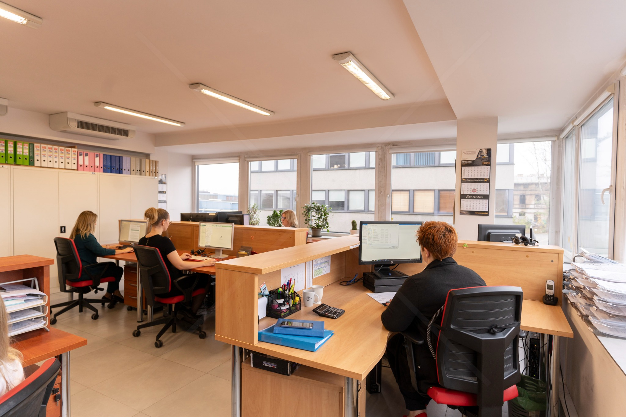 Jasne biuro z pracownikami przy biurkach, monitory, segregatory, kalendarz na ścianie. Widok na okna i budynki za nimi. Praca biurowa w nowoczesnym wnętrzu.