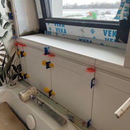 Firma Handlowo Usługowa Volodymyr Tsymbal - Białe płytki ścienne z kolorowymi klipsami poziomującymi, zamontowane wzdłuż okna. Gumowy młotek leży na wannie. Widok na zimowy krajobraz za oknem.