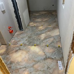 Firma Handlowo Usługowa Volodymyr Tsymbal - Układanie marmurowych płytek podłogowych z żółtymi klipsami poziomującymi w trakcie remontu. Widoczne rury i płyty gipsowe na ścianach.