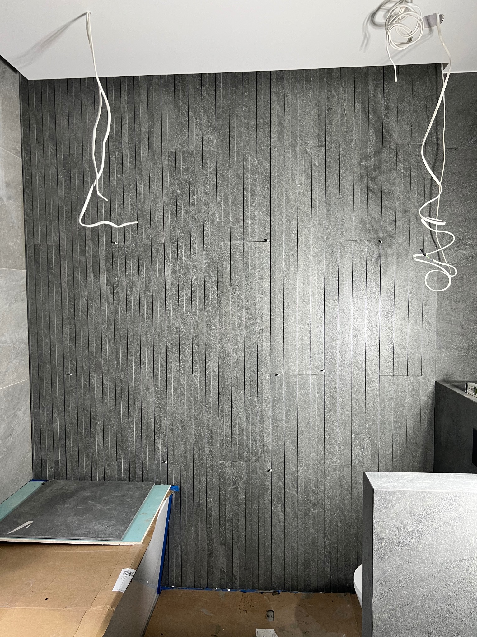 Ściana w łazience wykończona wąskimi, szarymi płytkami imitującymi kamień. Widoczne wystające przewody elektryczne i przygotowane płytki na podłodze.