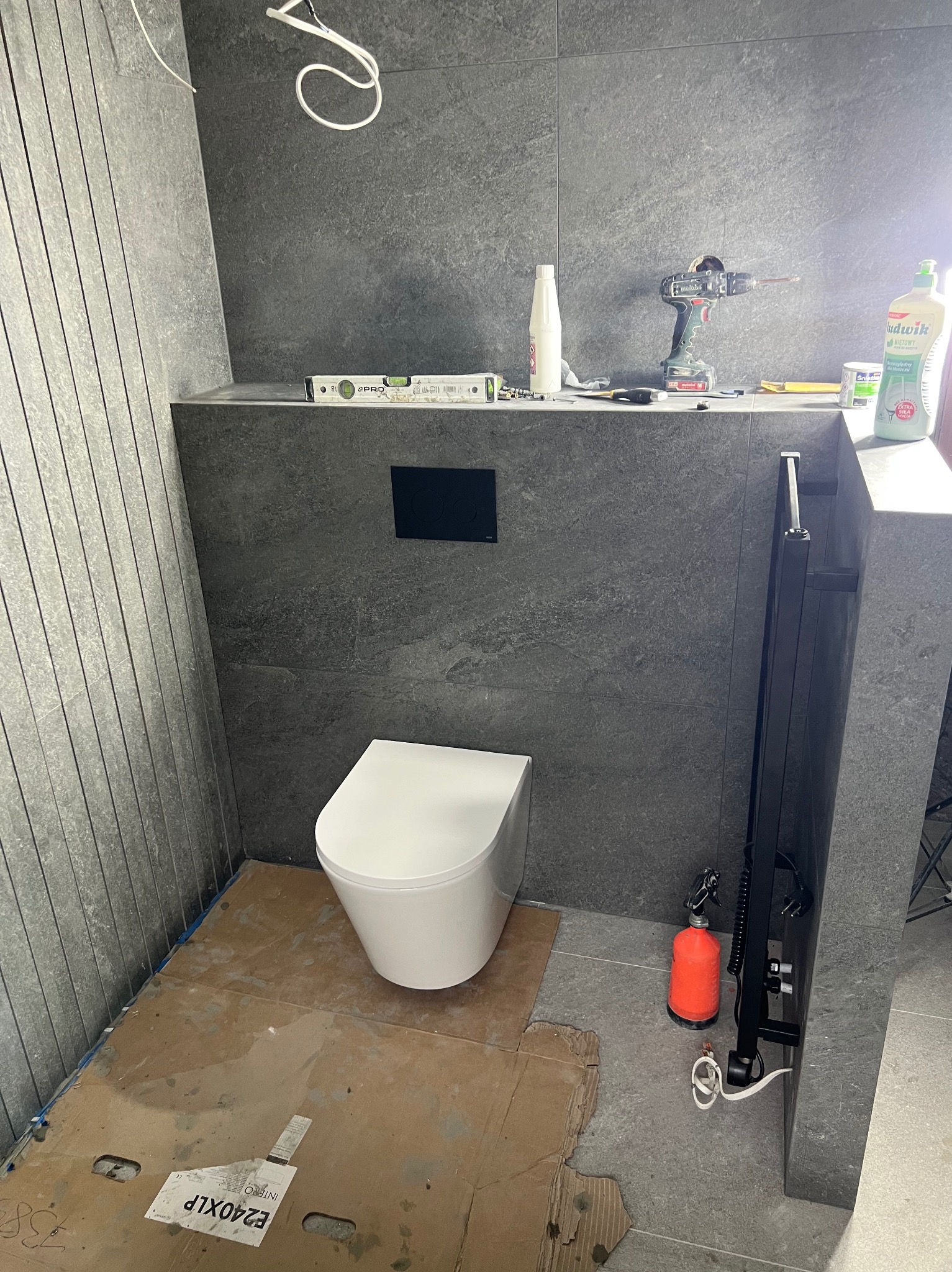 Wnętrze łazienki w trakcie remontu: widoczna toaleta, szare płytki ścienne, narzędzia i akcesoria glazurnicze. Ochronne kartony na podłodze.