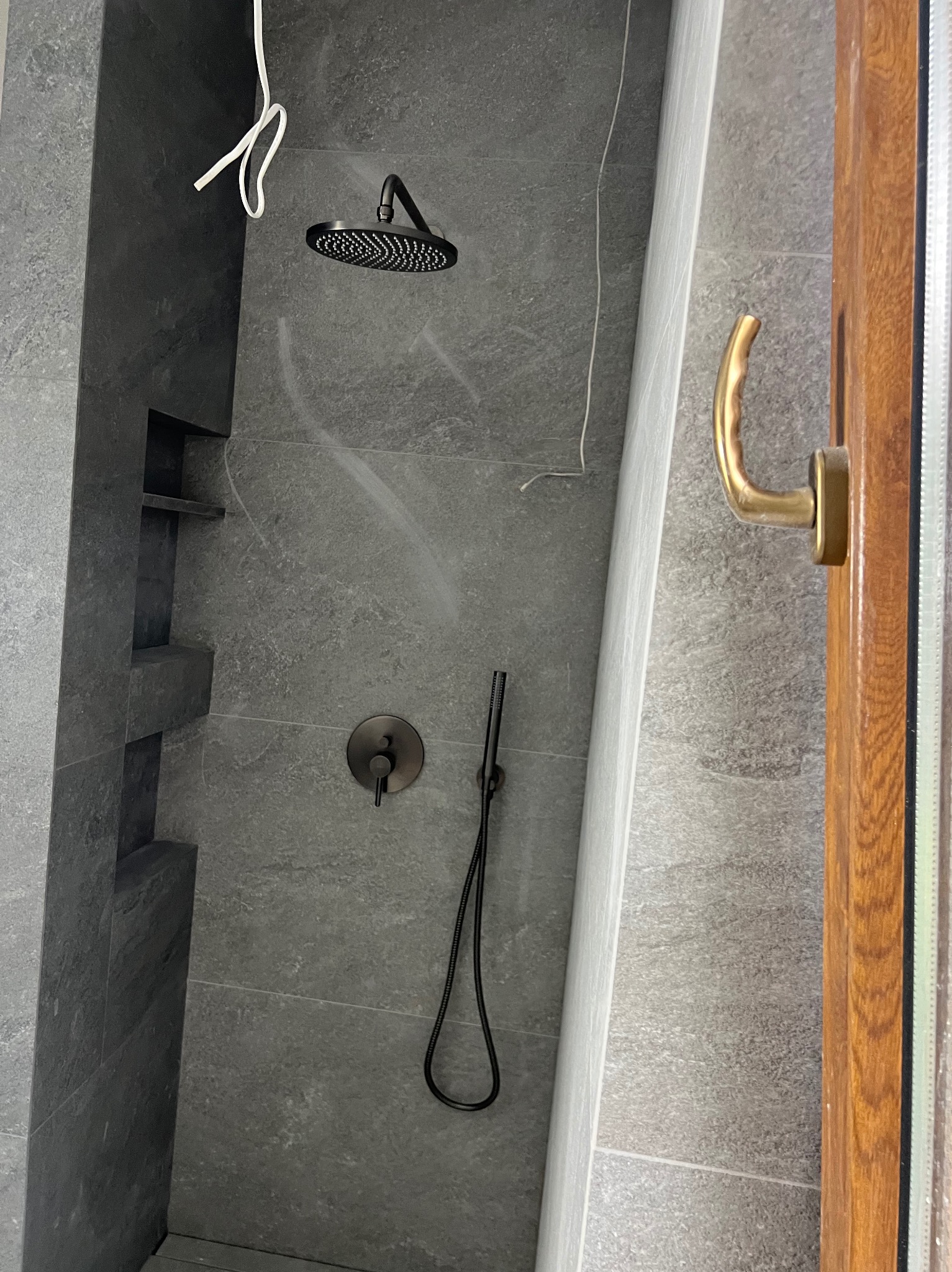 Wykończona kabina prysznicowa z szarymi płytkami, czarną armaturą i drewnianą ramą drzwi. Widoczne półki w ścianie. Minimalistyczny design.