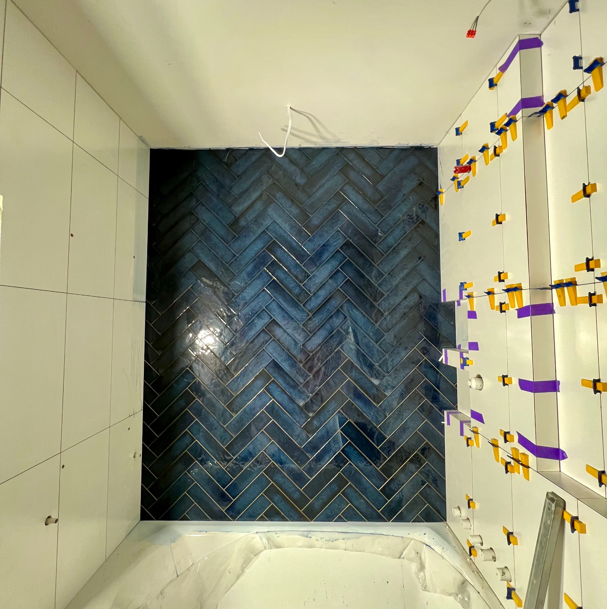 Łazienka w trakcie wykańczania: białe płytki na ścianach z żółtymi klipsami, fragment ściany z ciemnoniebieską glazurą ułożoną w jodełkę.