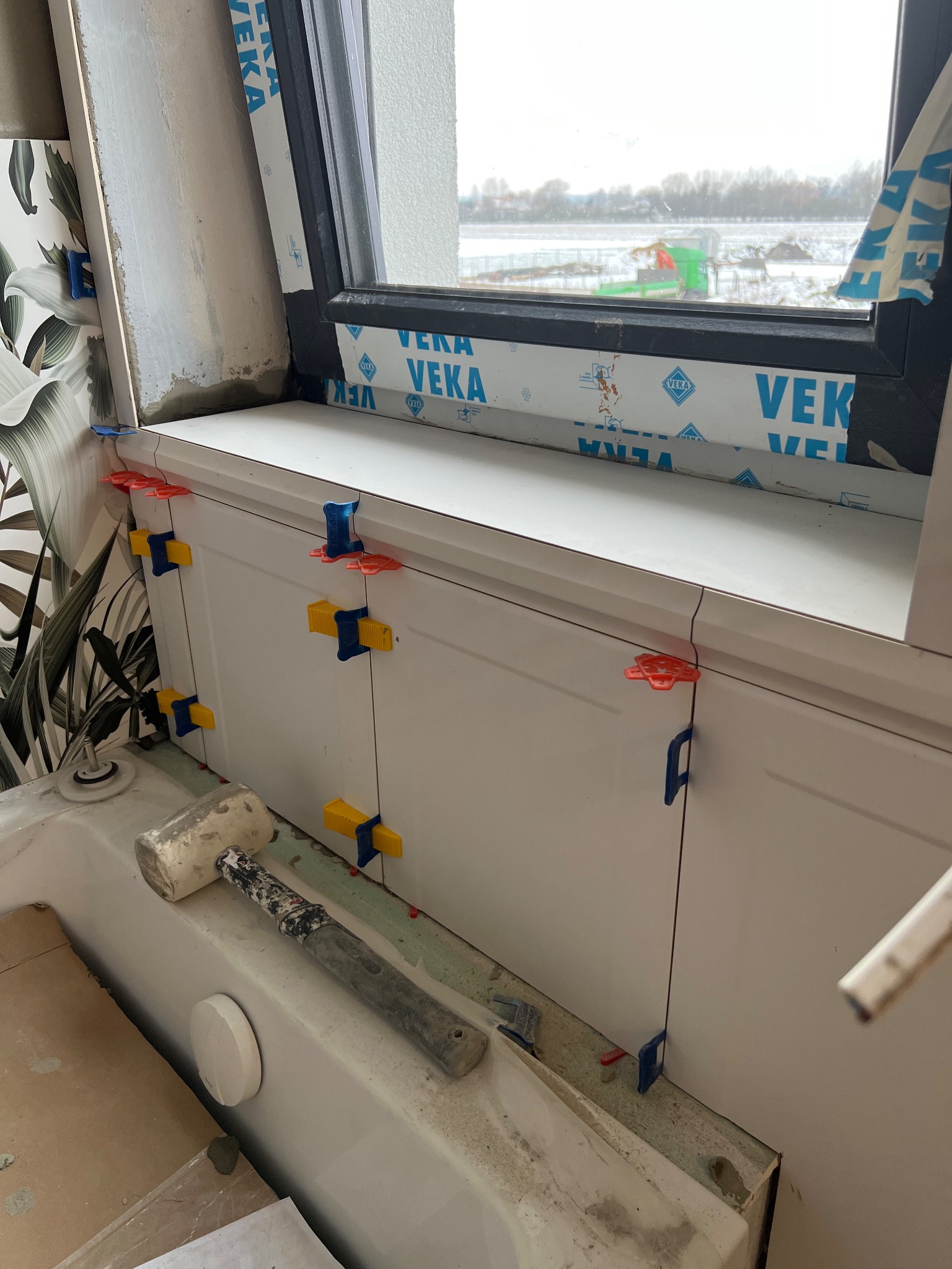 Białe płytki ścienne z kolorowymi klipsami poziomującymi, zamontowane wzdłuż okna. Gumowy młotek leży na wannie. Widok na zimowy krajobraz za oknem.