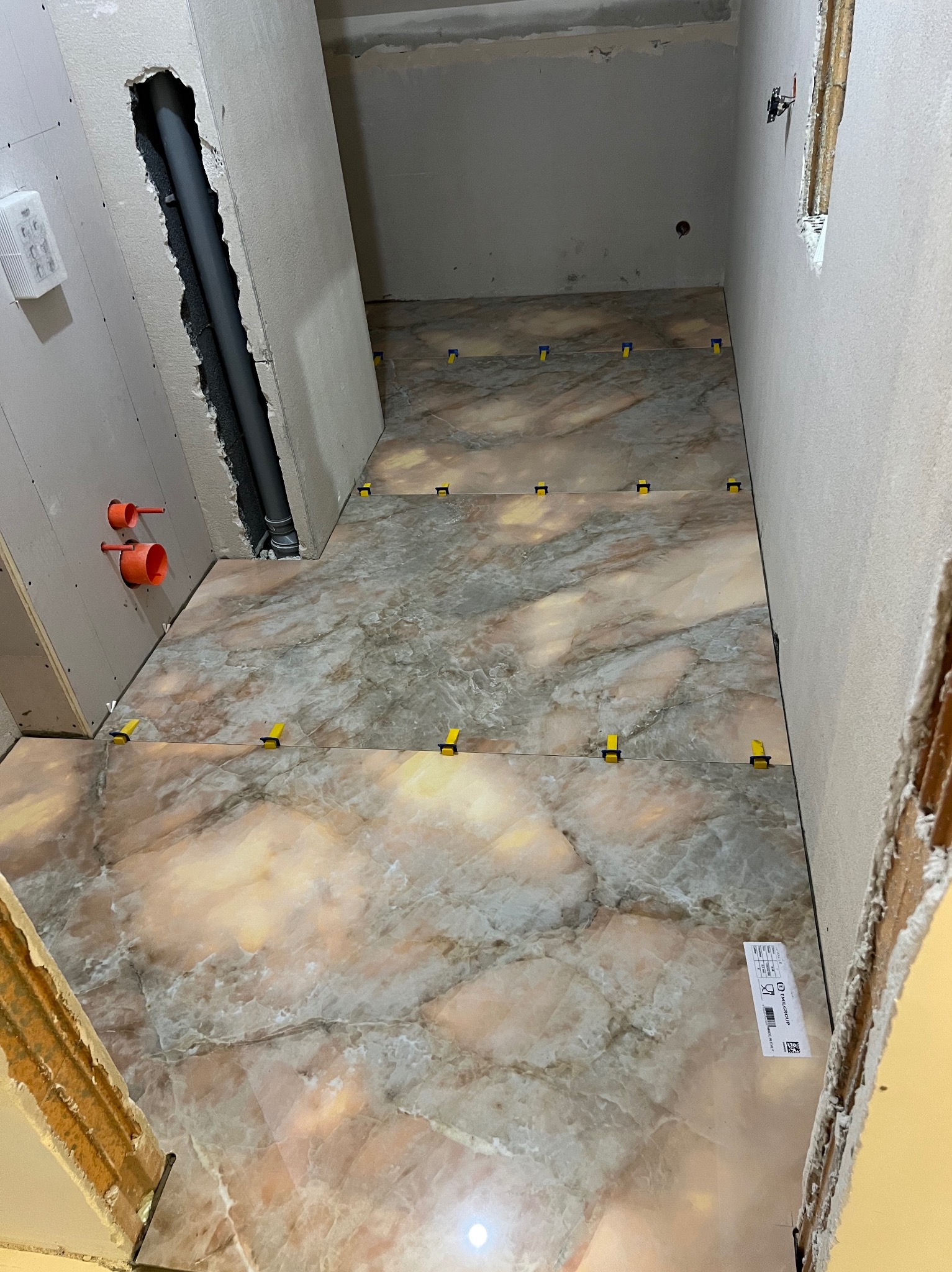 Układanie marmurowych płytek podłogowych z żółtymi klipsami poziomującymi w trakcie remontu. Widoczne rury i płyty gipsowe na ścianach.