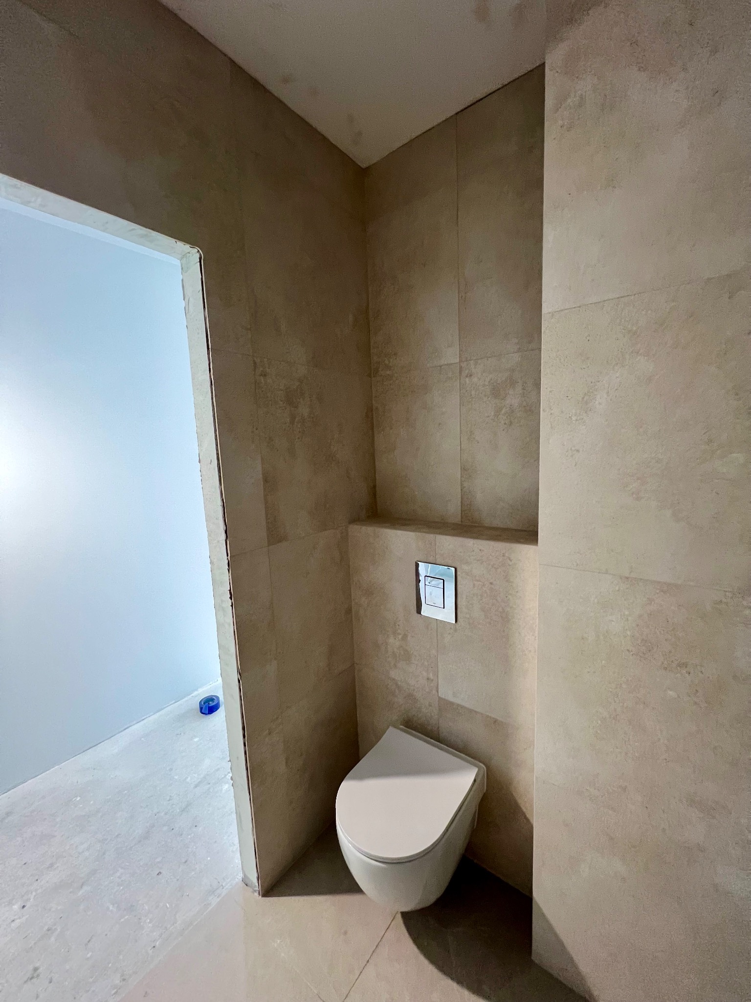 Beżowa łazienka w trakcie wykańczania: podwieszana toaleta, płytki imitujące kamień, widoczne niedokończone obramowanie drzwi. Minimalistyczny styl.