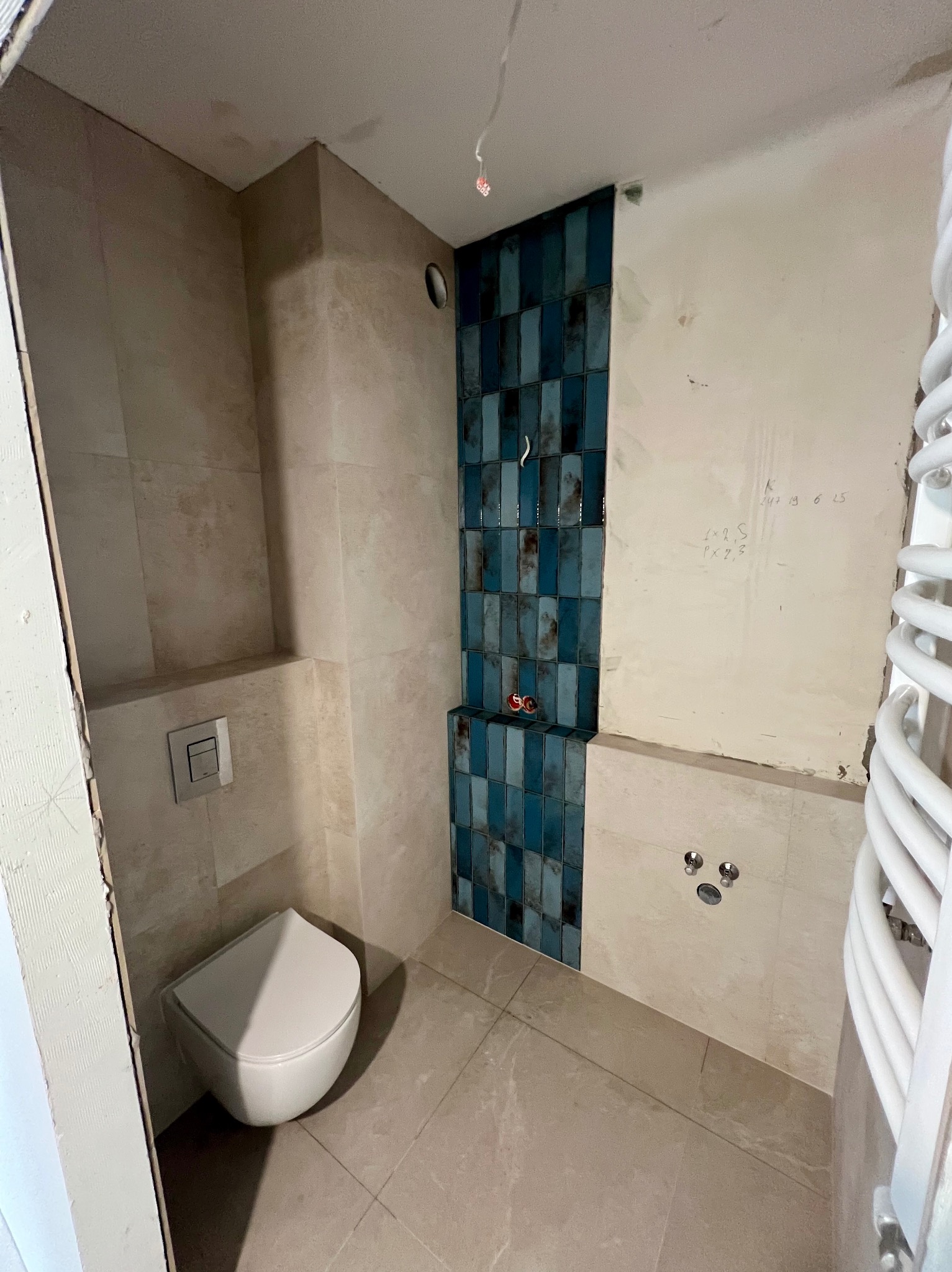 Wnętrze łazienki w trakcie wykańczania: beżowe płytki, fragment ściany z niebieską mozaiką, podwieszana toaleta, grzejnik drabinkowy i instalacja pod armaturę.