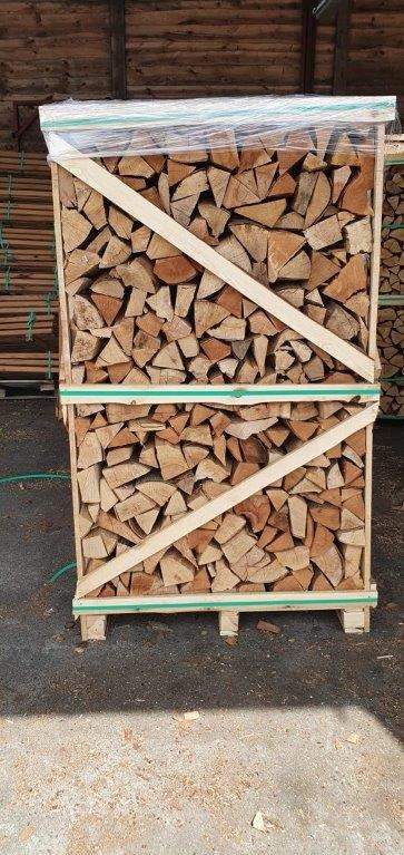 Paleta szczap drewna opałowego, zabezpieczona folią i drewnianą ramą z zielonymi paskami, na tle ściany z desek.