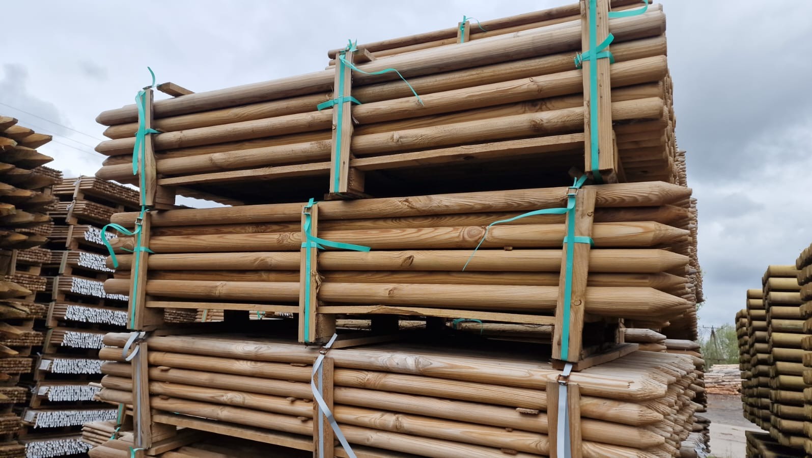 Stosy impregnowanych, zaostrzonych drewnianych słupków na paletach, zabezpieczonych niebieską taśmą, składowane na zewnątrz przy pochmurnym niebie.
