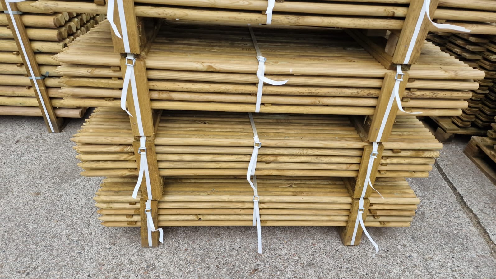 Stosy impregnowanych, ostro zakończonych drewnianych słupków ogrodzeniowych spięte białymi taśmami na paletach, gotowe do transportu.