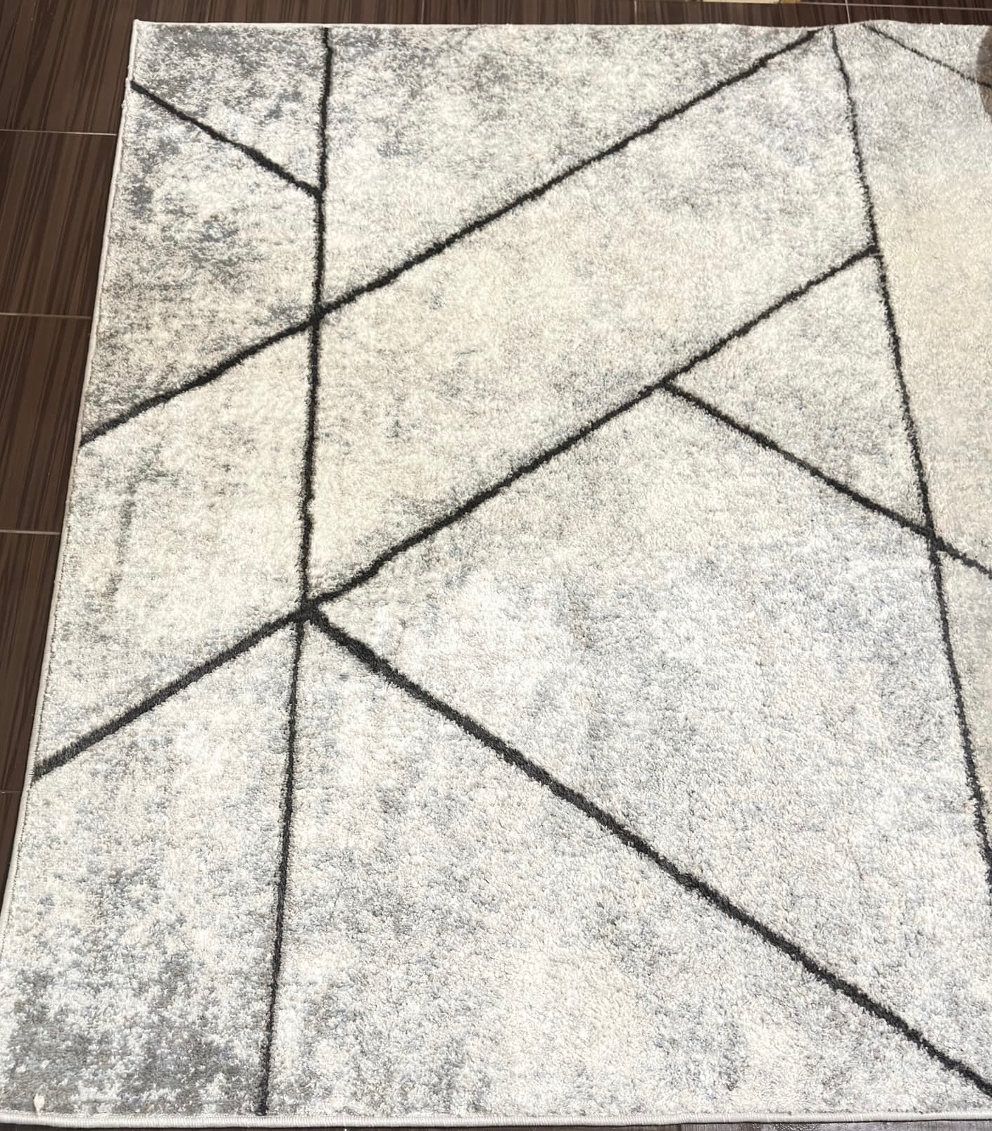 Jasnoszary dywan z geometrycznym wzorem z ciemnych linii na brązowej podłodze.