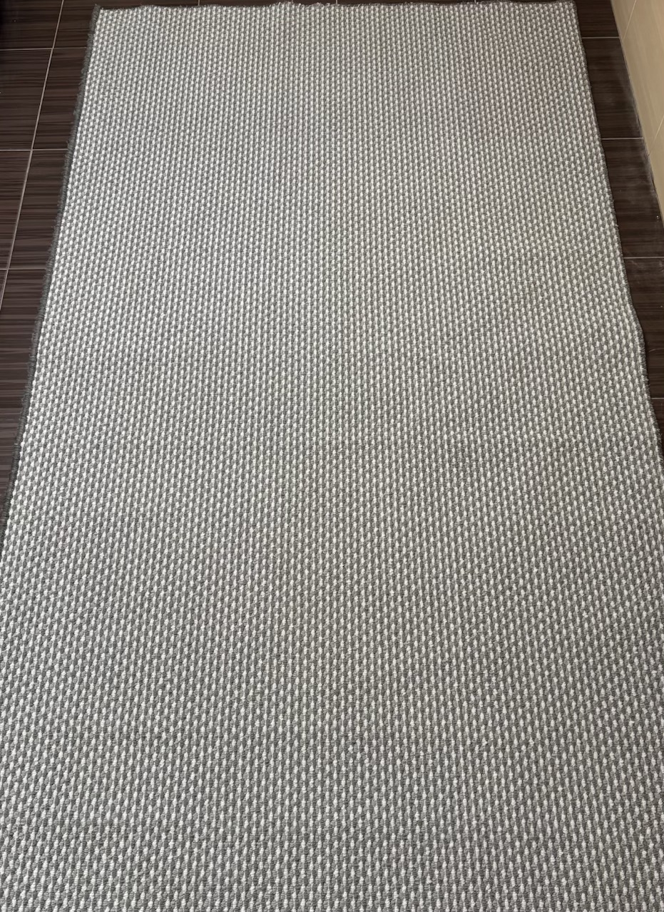 Szary dywan o drobnym, prążkowanym wzorze leżący na brązowych płytkach podłogowych.