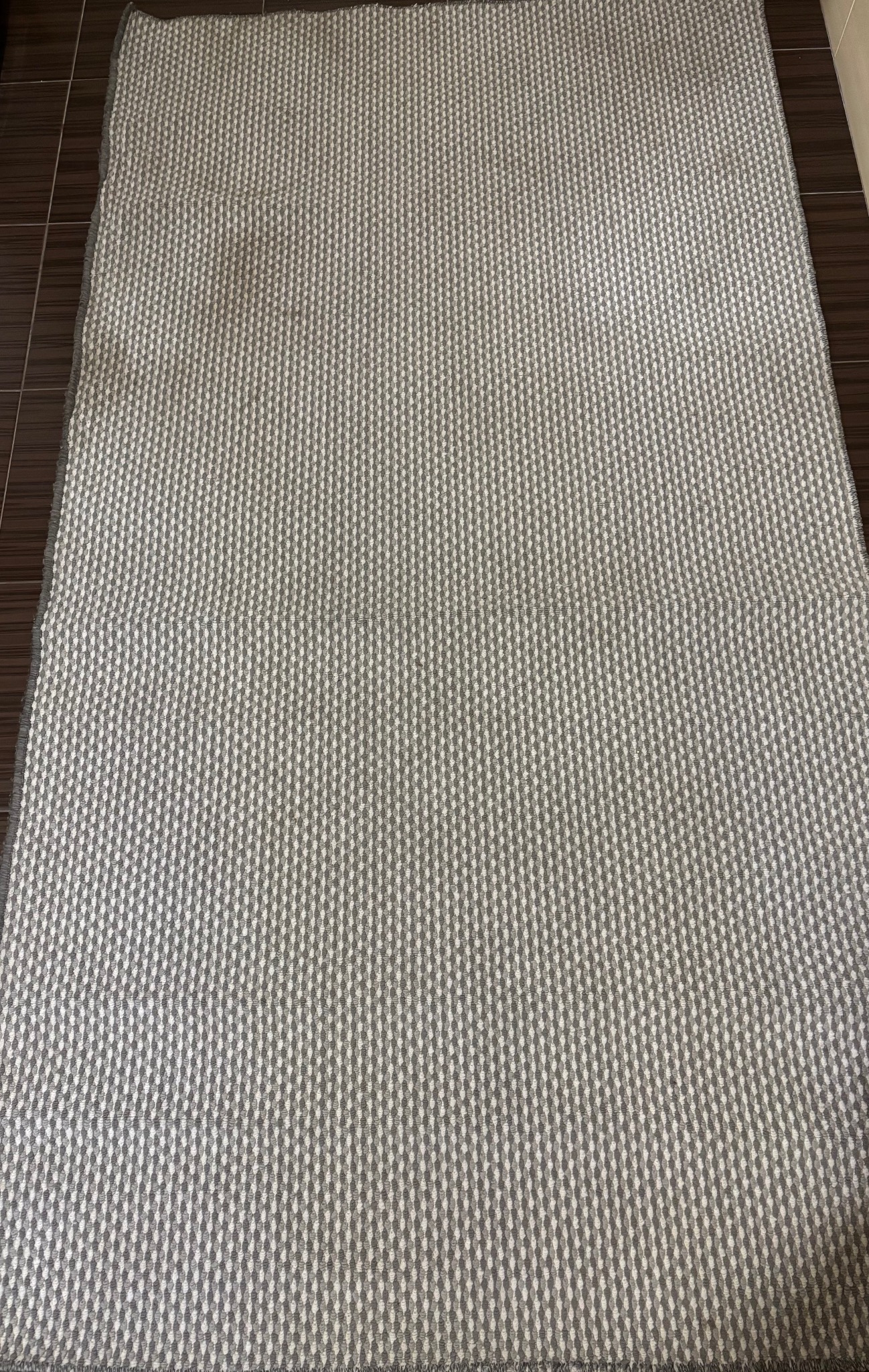 Szaro-biały dywan z geometrycznym wzorem w pionowe prążki, leżący na ciemnobrązowej podłodze z płytek ceramicznych.