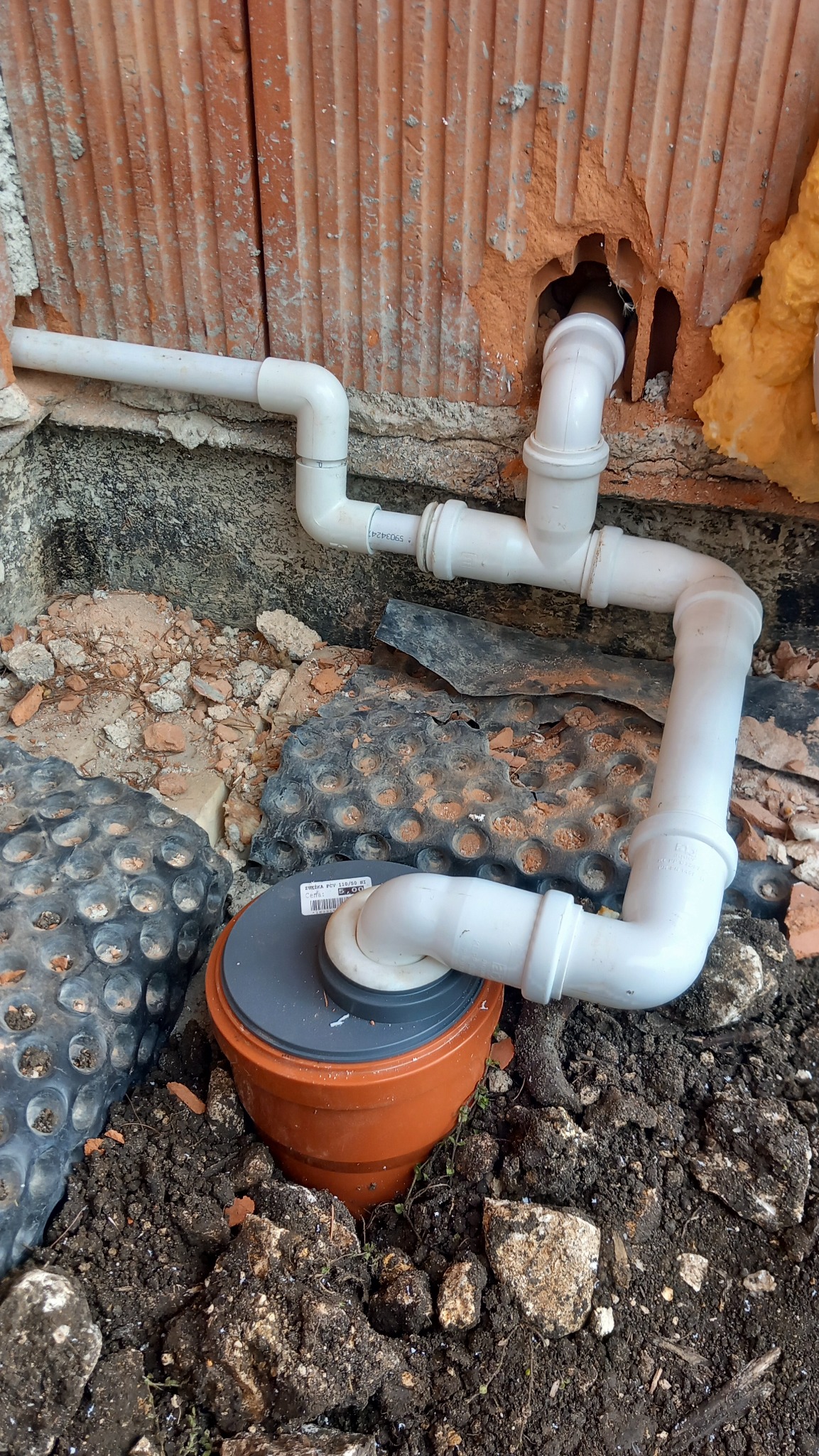 Instalacja odpływowa z białych rur PCV, wkopana w ziemię przy ceglanej ścianie z widoczną pianką montażową. Elementy instalacji połączone pod kątem.