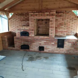 Murowany piec z cegły z trzema otworami, w tym dwa z żeliwnymi drzwiczkami, pod drewnianą konstrukcją zadaszenia, na szarym brukowanym podłożu z widocznymi śladami wody.