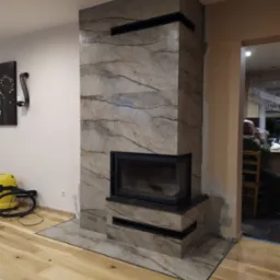 Obudowa kominkowa z marmurowym wzorem, czarny wkład kominkowy, drewniana podłoga, widoczny odkurzacz i osoba w tle.