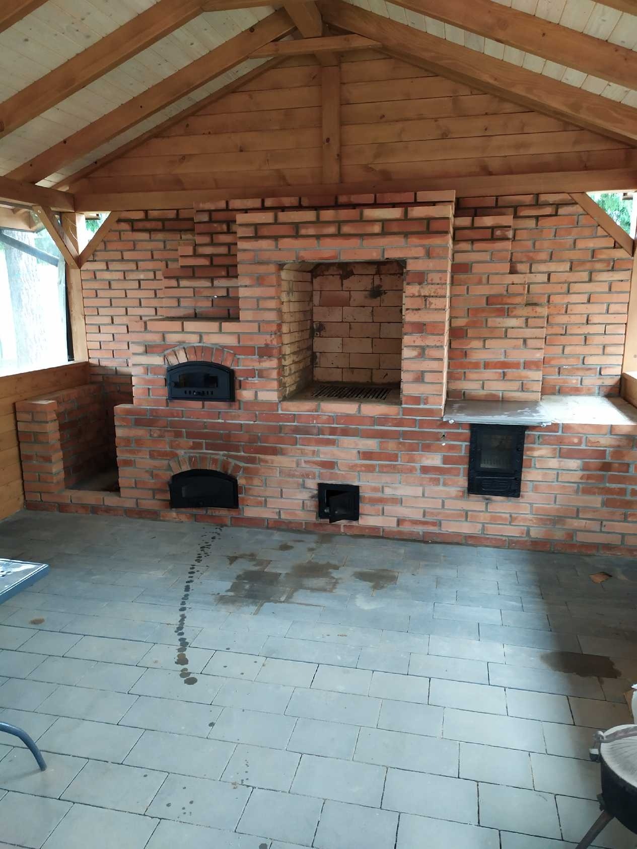 Murowany piec z cegły z trzema otworami, w tym dwa z żeliwnymi drzwiczkami, pod drewnianą konstrukcją zadaszenia, na szarym brukowanym podłożu z widocznymi śladami wody.