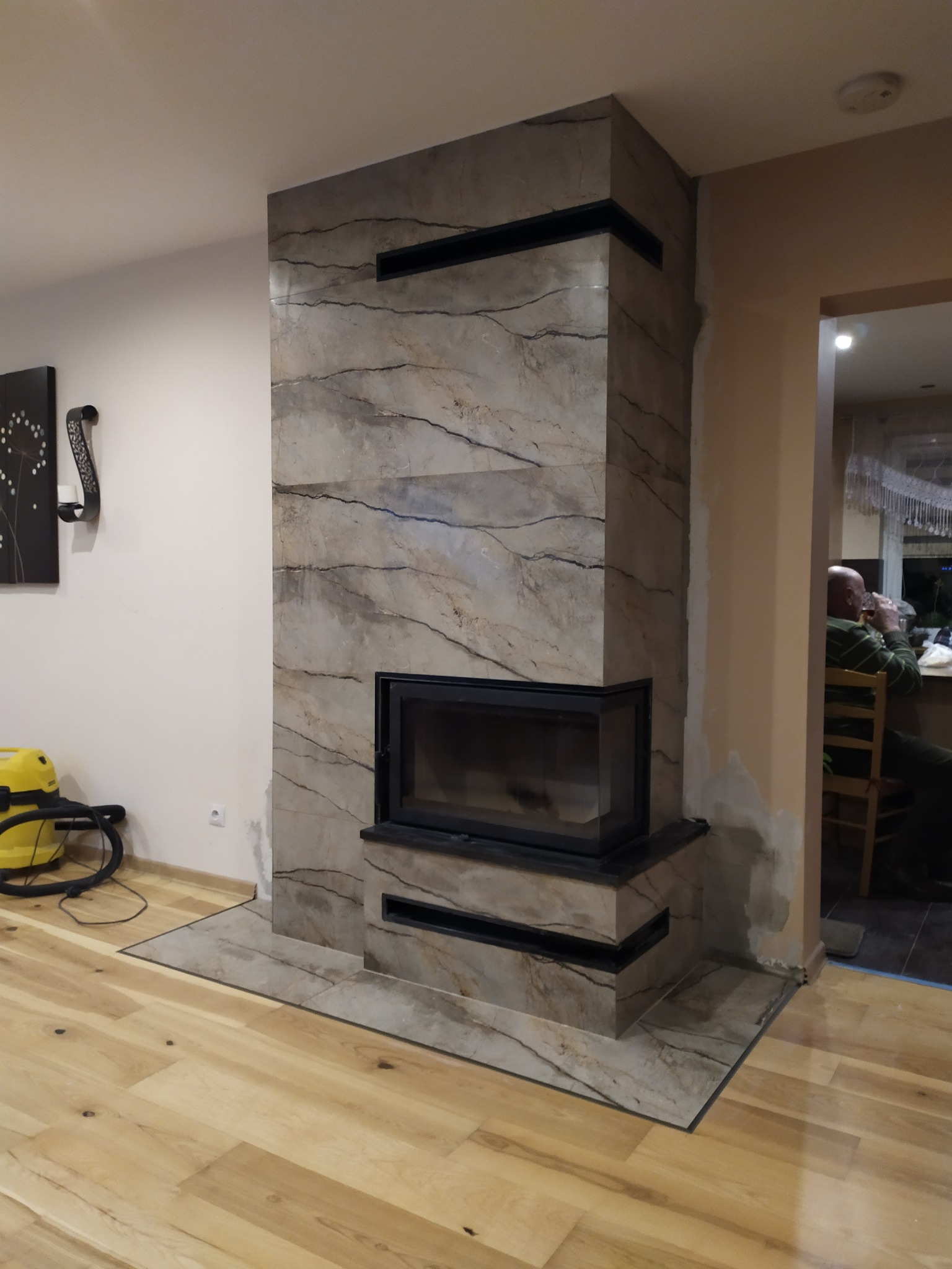 Obudowa kominkowa z marmurowym wzorem, czarny wkład kominkowy, drewniana podłoga, widoczny odkurzacz i osoba w tle.