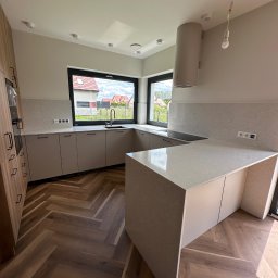 GlassStone Filip Wołoszuk - Jasna kuchnia z wyspą i blatem z delikatnym wzorem, widok na zewnątrz przez duże okno. Parkiet ułożony w jodełkę. Minimalistyczny design, nowoczesne oświetlenie.