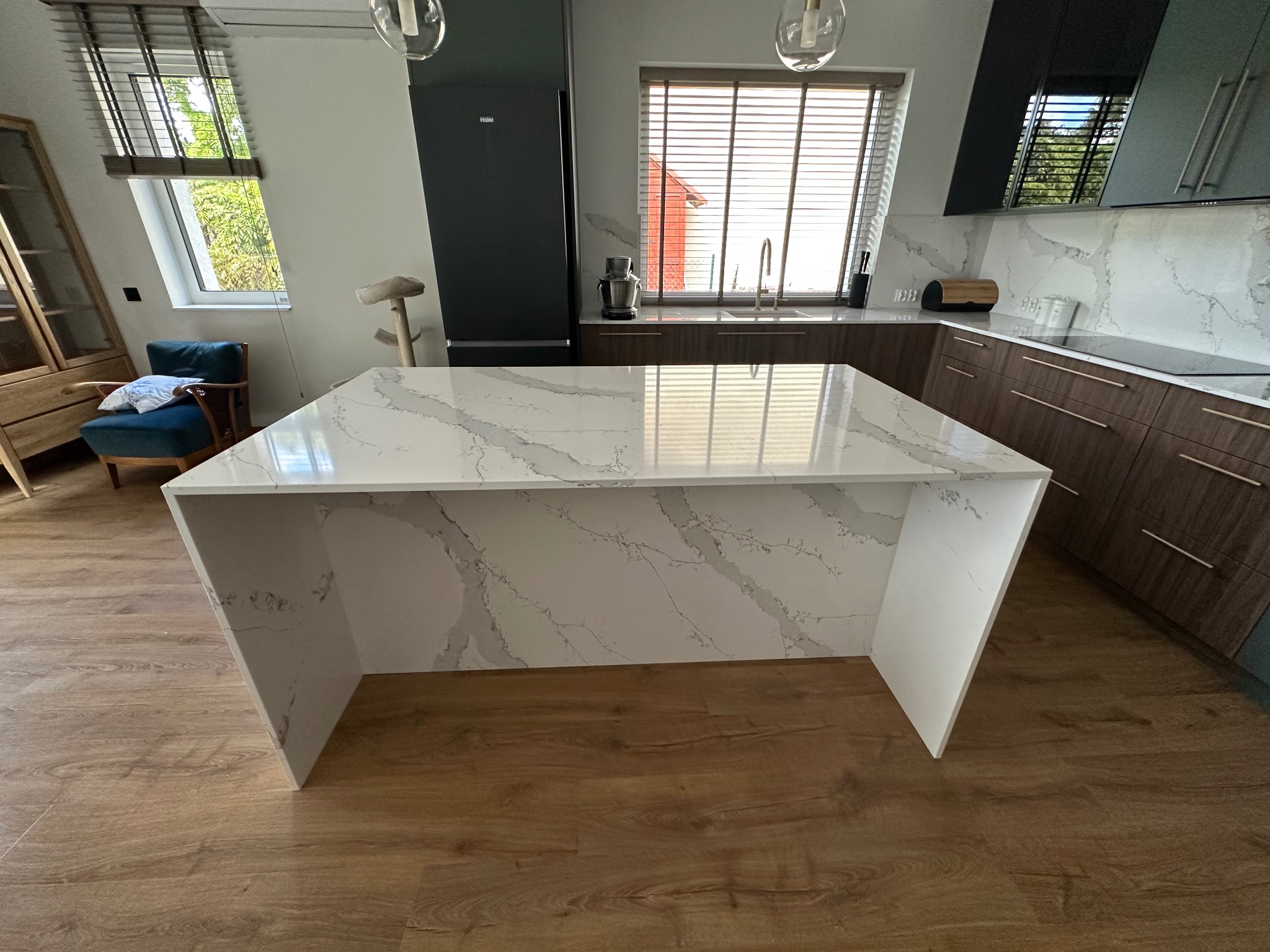 Kuchenna wyspa z blatem z białego kamienia z szarymi żyłami, na tle nowoczesnej kuchni z drewnianymi szafkami i oknem z żaluzjami. Widoczny fragment podłogi.