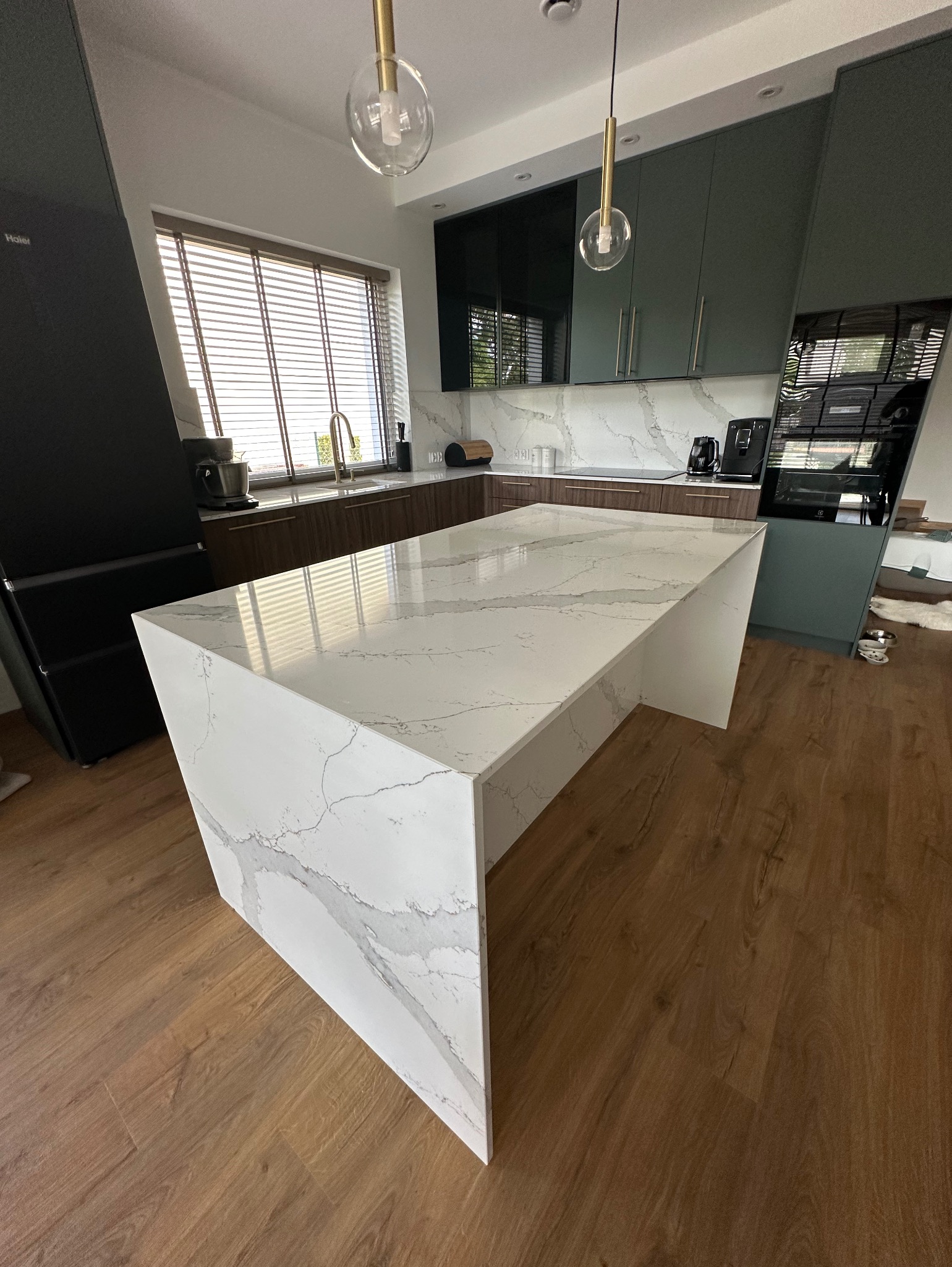 Biały blat kuchenny z marmurowym wzorem, oświetlony wiszącymi lampami, w nowoczesnej kuchni z drewnianą podłogą i oknem z żaluzjami.