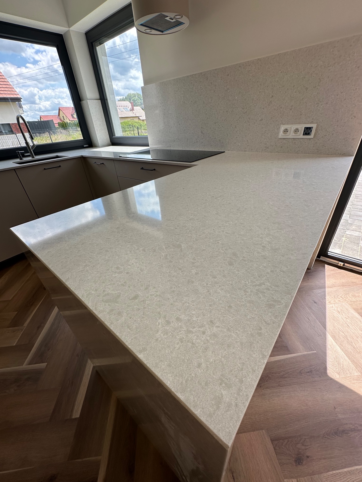 Jasny blat kamienny w nowoczesnej kuchni z oknem i parkietem w jodełkę. Widoczne gniazdka elektryczne na ścianie i fragment zlewu. Blat oświetlony naturalnym światłem.