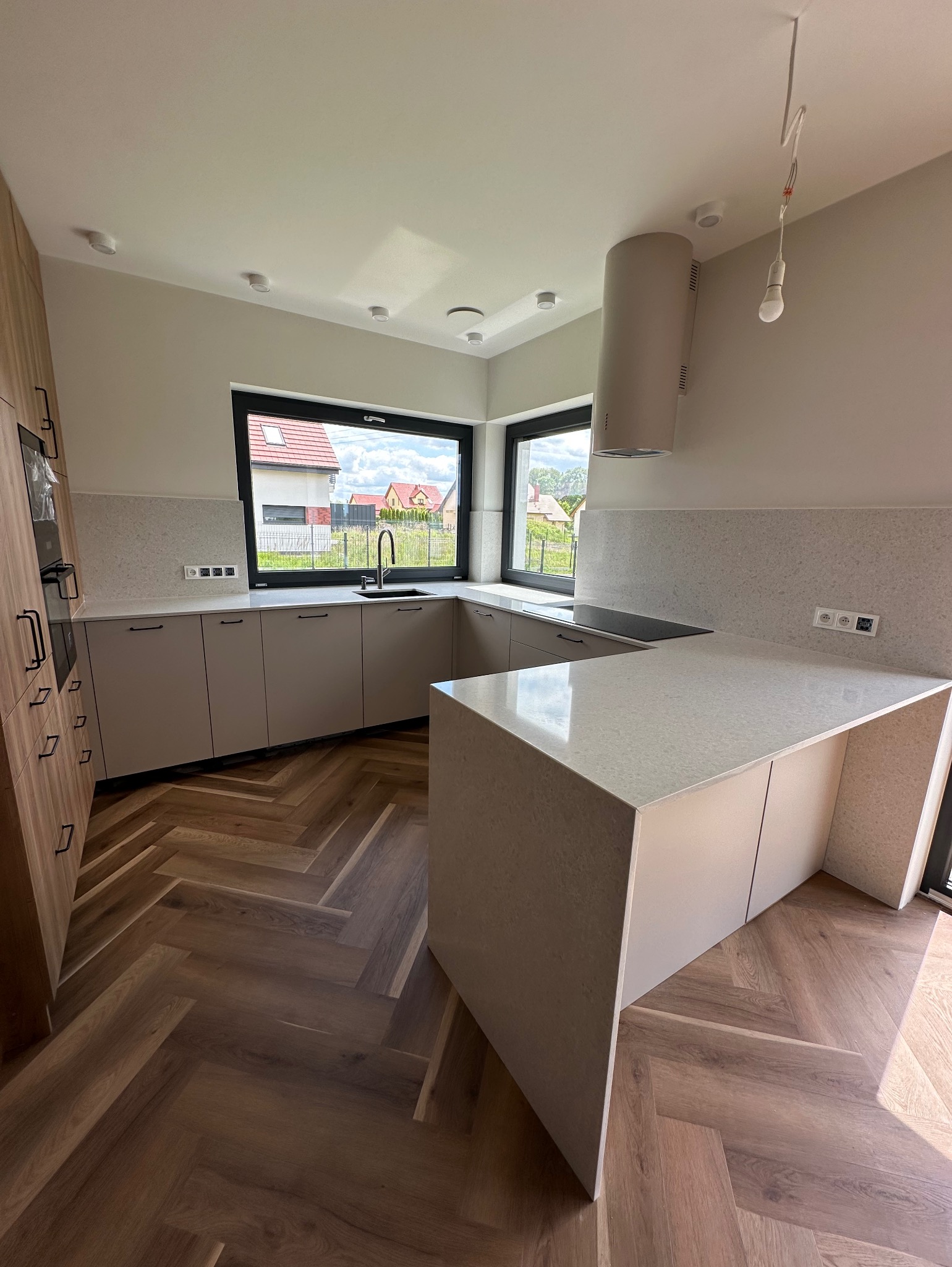 Jasna kuchnia z wyspą i blatem z delikatnym wzorem, widok na zewnątrz przez duże okno. Parkiet ułożony w jodełkę. Minimalistyczny design, nowoczesne oświetlenie.