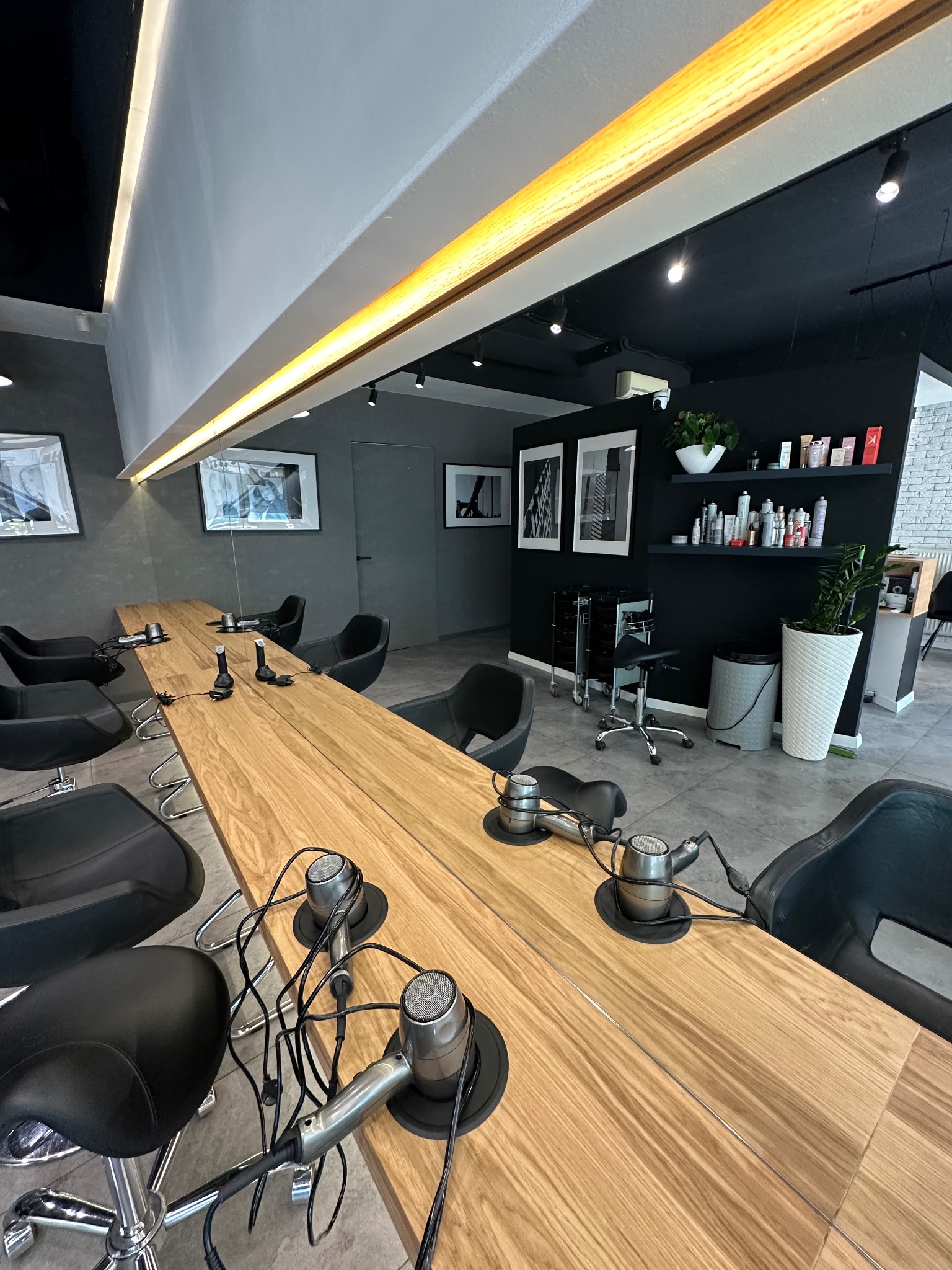 Nowoczesne wnętrze salonu fryzjerskiego z długim, drewnianym blatem z wbudowanymi suszarkami, czarnymi krzesłami i minimalistycznym wystrojem w odcieniach szarości i czerni.