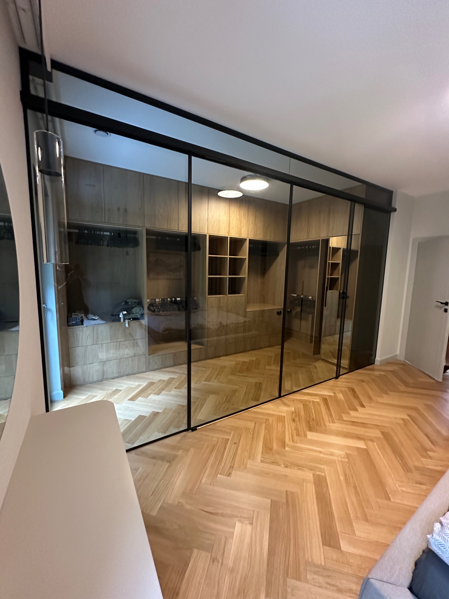 Nowoczesna garderoba za przesuwnymi drzwiami ze szkła w czarnej ramie, parkiet w jodełkę, oświetlenie sufitowe. Widok na wnętrze szafy z półkami i wieszakami.