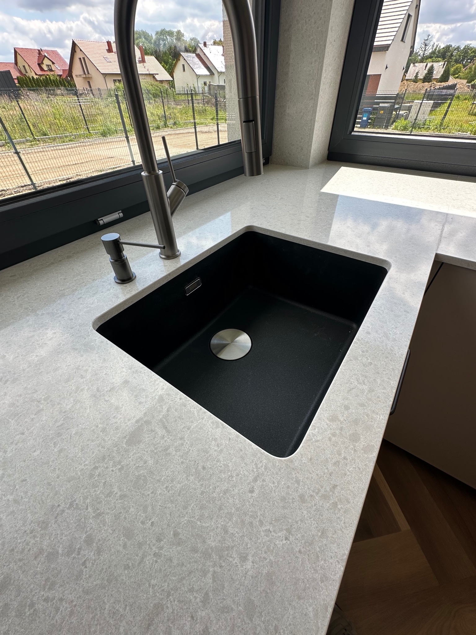 Jasnoszary blat z kamienia kwarcowego z czarnym zlewem i baterią w nowoczesnej kuchni, widok z góry, z oknem w tle.