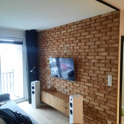 PŁOTKA REMONTY I WYKOŃCZENIA SZYMON PŁOTKA - Salon z dekoracyjną ścianą z cegły, telewizorem, drewnianą szafką RTV i białymi głośnikami podświetlanymi przez lampę wiszącą przy balkonie.