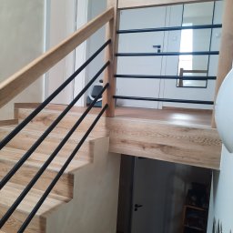 PŁOTKA REMONTY I WYKOŃCZENIA SZYMON PŁOTKA - Nowoczesne schody drewniane z czarnymi, metalowymi balustradami, wykończone płytkami imitującymi drewno, widok z dołu na górny poziom z białymi drzwiami i lustrem.
