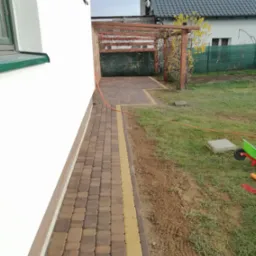 Ułożona kostka brukowa w dwóch odcieniach brązu i żółci przy ścianie budynku w Welk, widoczna pergola i trawnik.