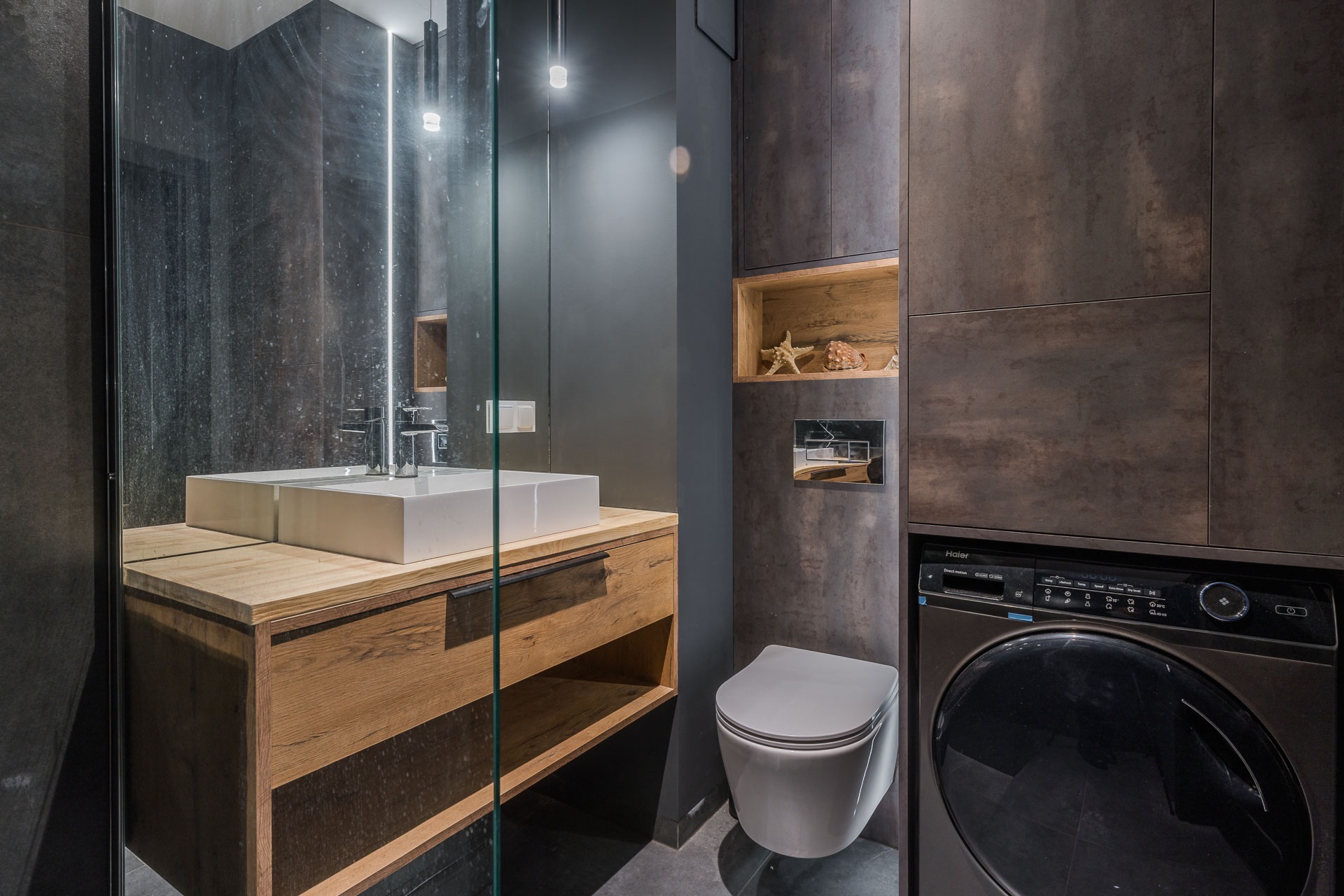 Nowoczesna łazienka w odcieniach szarości z drewnianą szafką pod umywalką, kabiną prysznicową ze szklaną ścianką, podwieszaną toaletą i pralką w zabudowie, uzupełniona o półkę z dekoracjami morskimi.