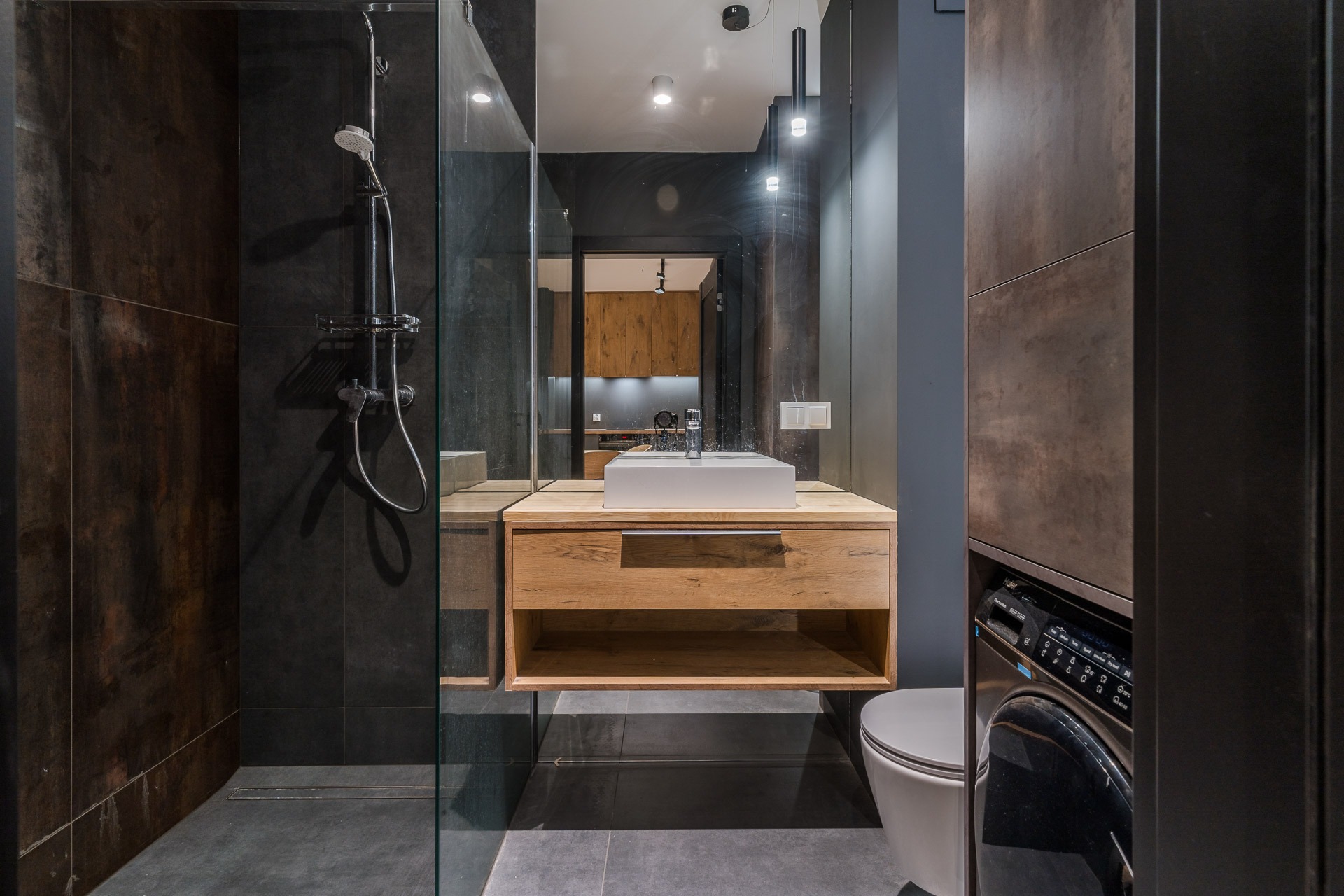 Nowoczesna łazienka z kabiną prysznicową typu walk-in, umywalką nablatową na drewnianej szafce, toaletą oraz pralką ukrytą w zabudowie; ciemne płytki imitujące metal na ścianach i podłodze...